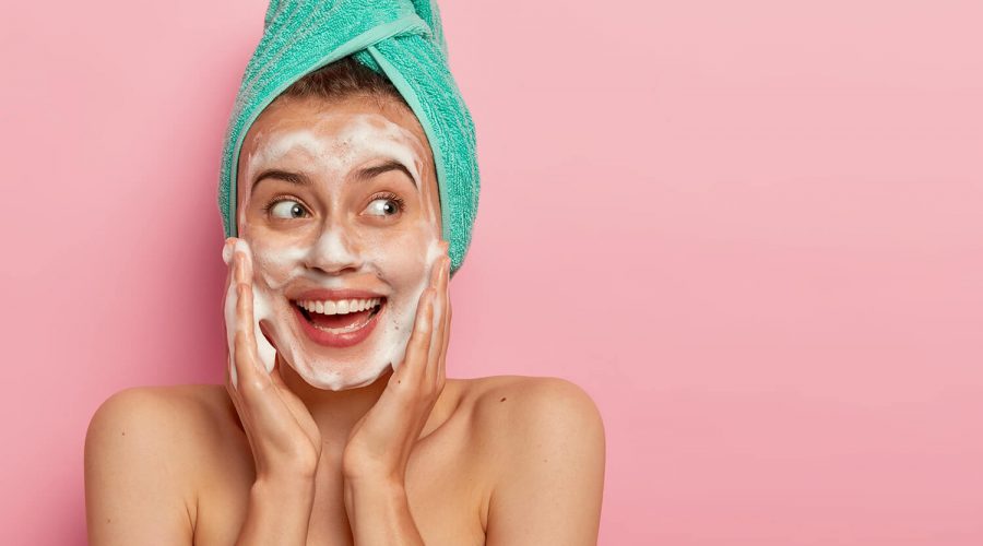 headshot-of-lovely-smiling-female-model-touches-cheeks-looks-aside-washes-face-with-soap-bubble-has-naked-body-wears-turquoise-soft-towel-on-head-poses-over-rosy-wall-copy-space-for-promotion (1)